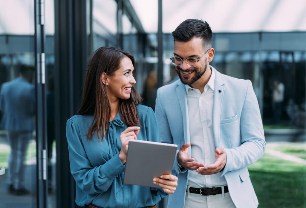 Shot of two confident colleagues walking and talking next to an office building. Businessman and businesswoman in meeting using digital tablet and discussing business strategy. Creative business persons discussing new project and sharing ideas while walking in front of their office building.