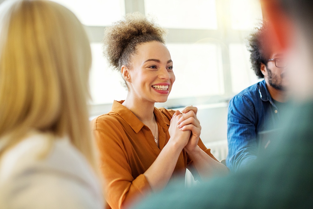 Happy businesswoman with hands clasped looking away. She is sitting with colleagues in office. Focus is on her.